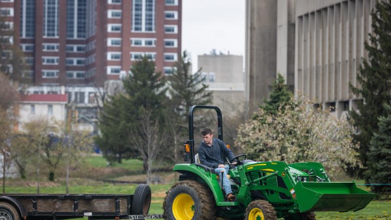 Student driving a John Deere tractor with a wagon attached in front of the Gluck building during a UK Field Day competition